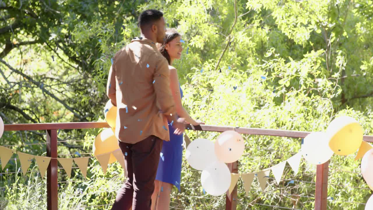 Couple walking onto deck past balloons pausing at railing man placing hand taking view during party