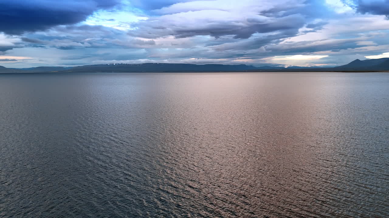 Smooth surface of the sea or ocean reflecting grey clouds. Aerial perspective on the waterscape with mountain range at backdrop.