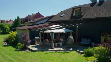 Toma-Aérea-De-Drones-Del-Jardín-Y-Terraza-Con-Muebles-Junto-A-La-Casa-Familiar-Durante-El-Verano