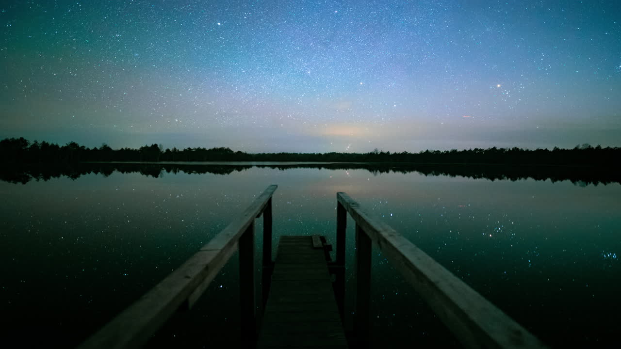 Wooden dock leads out to lake reflecting mesmerizing starry night sky in forest