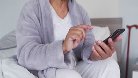 sección media de una mujer mayor afroamericana que usa un teléfono inteligente mientras está sentada en la cama en casa
