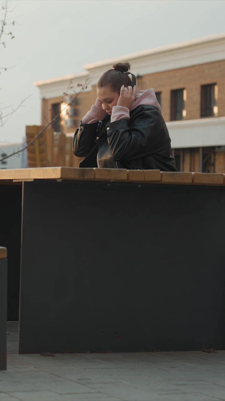 Soggetto con capelli scuri raccolti che indossa una giacca di pelle. Il soggetto solleva una fascia verso il viso preparandosi all'uso. La scena urbana esterna presenta edifici in mattoni, alberi spogli, strutture in legno.
