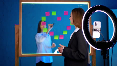 two women are shooting video for blog indoors. bloggers talk in front of phone camera with light ring displaying glass wall with sticky notes.