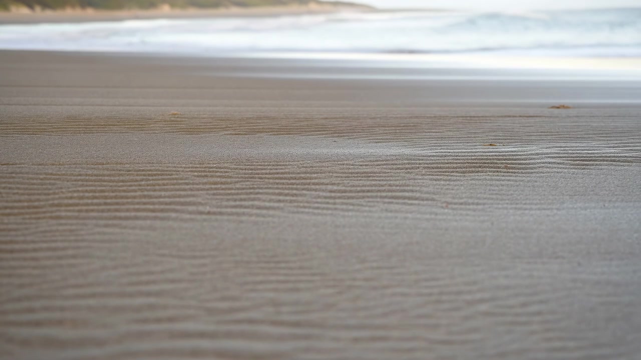 Lavvinklet kystudsigt, der viser teksturerede våde sandkrusninger i forgrunden med lav dybdeskarphed, blød bokeh i mellemgrunden, fjern kystlinje og blid.