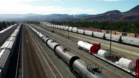 truck-left-drone-shot-flying-over-railroad-station-in-a-desert-environment-on-a-sunny-day-over-black-tank-trains-and-other-cargo-trains