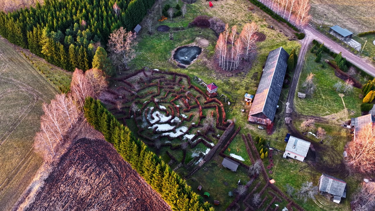 Labyrinth hedge maze with central pond and farm buildings during spring