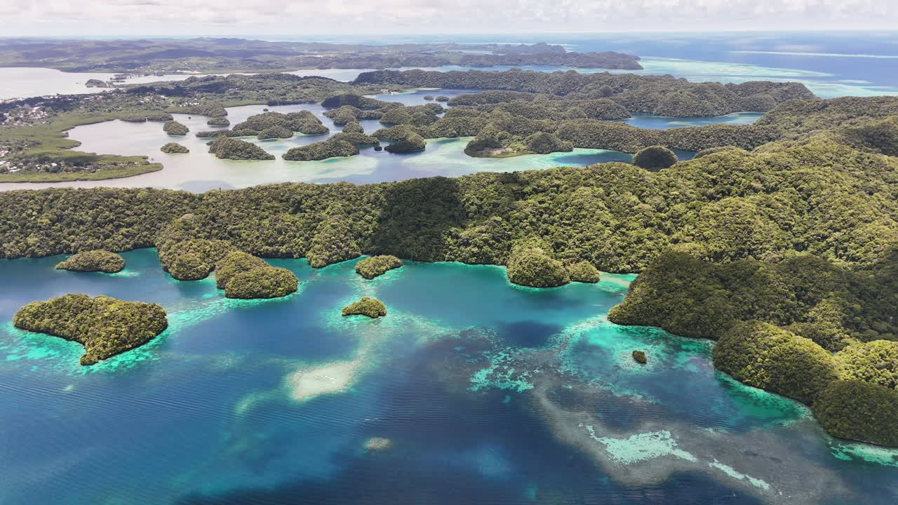 Scenic drone shot sweeping across the vivid aqua ocean and green-covered terrain with a small town in the distance in Palau. Wide shot of town and islands