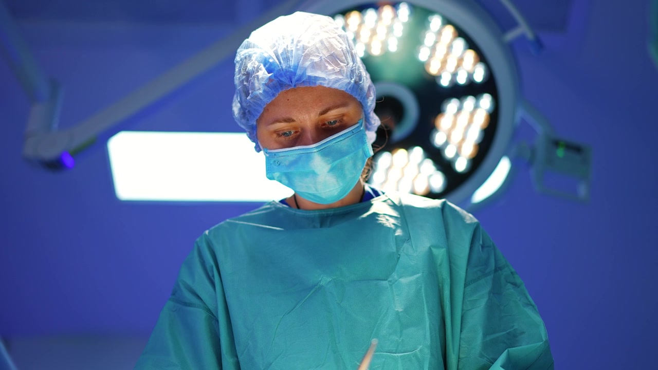 Una chirurga con cuffia, mascherina e camice opera. Vista dal basso del medico che esegue l'intervento. Una lampada rotonda sullo sfondo.