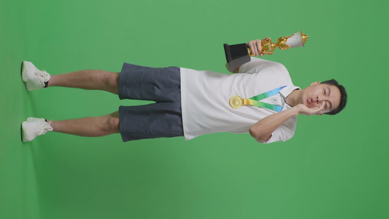 황금 메달을 들고 있는 아시아 남자의 몸이 가득 차 있고, 황금 트로피를 들고 있으며, 스튜디오의 녹색 스크린 배경에서 입을 고 손으로 긍정적으로 소리치고 있다.