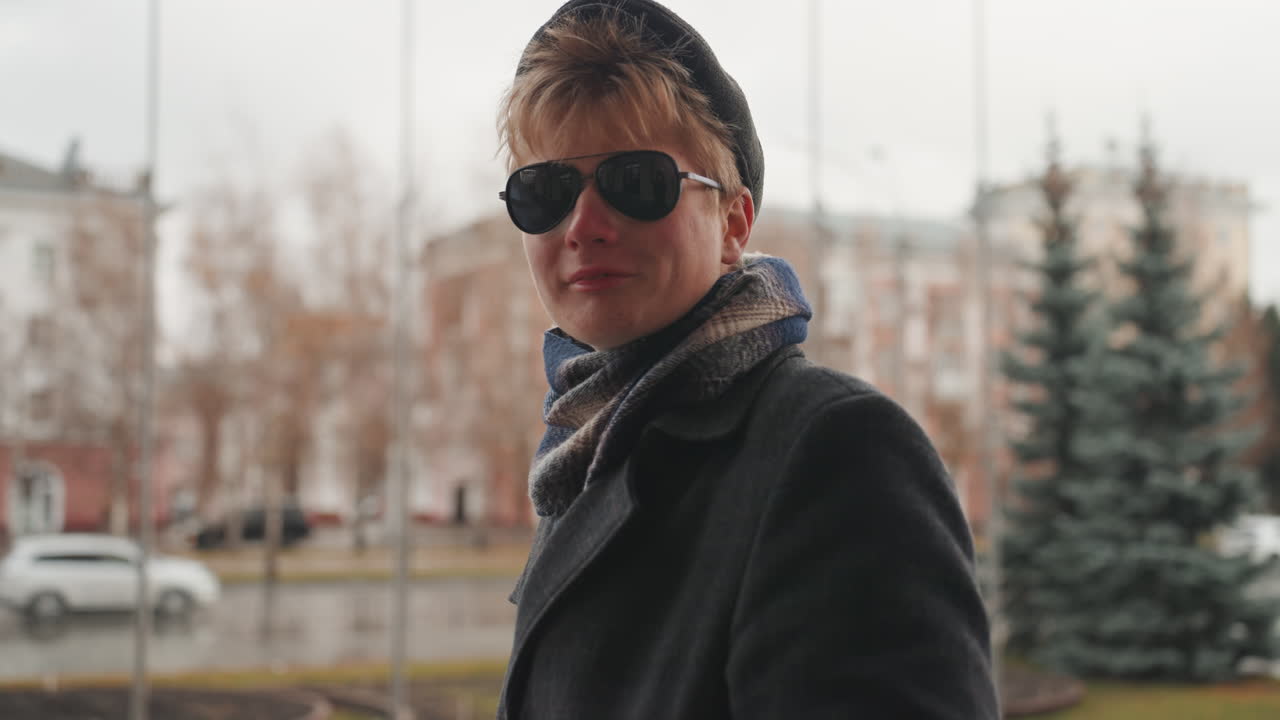 White creator stands in streetwear pose, confident portrait of white man in coat, scarf, sunglasses and beanie, rainslicked city background with pine trees and passing cars, editorial aesthetic