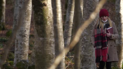 woman walking alone among trees in beech forest with book in hand