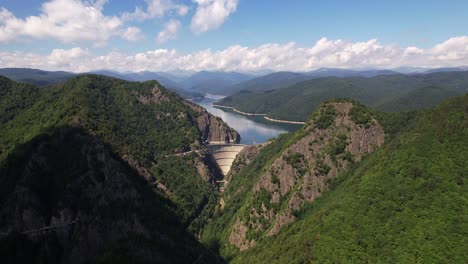 Vidraru-dam-in-romania,-with-the-lake-in-full-view,-surrounded-by-forested-mountains,-on-a-sunny-day,-aerial-view