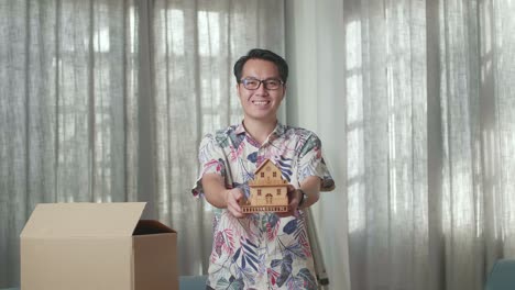 hombre asiático con cajas de cartón sonriendo y mostrando el modelo de casa a la cámara en la nueva casa
