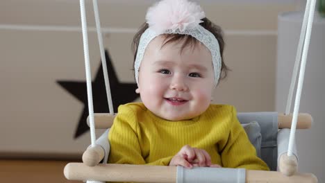 Hermosa-Sonrisa-Con-Dientes-De-Bebé-Mirando-A-La-Cámara-Mientras-Cuelga-En-Un-Columpio-Interior-En-Casa-Usando-Arco