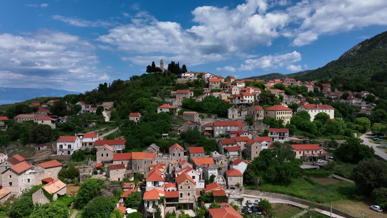 Hillside village with red-roofed houses under fluffy clouds against a bright blue sky