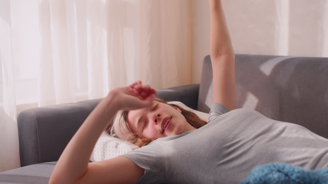 Lady finally wakes up and stretches herself on sofa, resting on white pillow with soft blanket, sunlight casting warm shadows across cozy living room