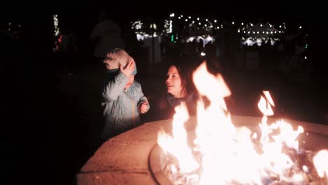 mother and three-year old toddler daughter at a firepit at night with christmas lights in the background