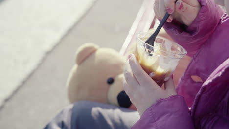 Close-up-of-little-girl-with-blue-jumper-nail-polish-and-teddy-bear-beside-her-holding-a-sundae-ice-cream-sitting-outdoor-4k