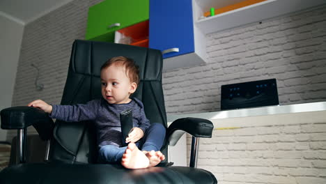 sweet baby boy sits in the office chair holding a tv remote. kid slaps happily the chair handrail with his hand. low angle view.
