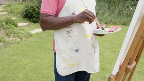 Mid-section-of-senior-african-american-man-painting-on-wooden-easel-in-garden,-slow-motion