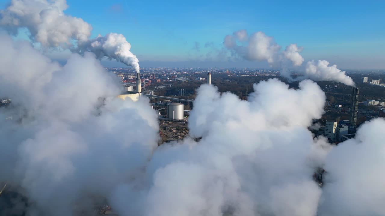 Aerial view of a thermal power plant emitting smoke on a sunny day, with a river and a city in the background. Great aerial view flight descending drone