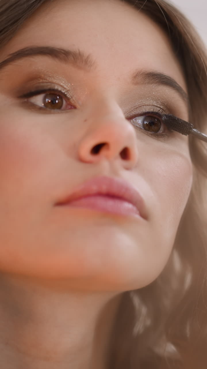 Focused woman applies mascara on long lashes to accentuate look. Curly-haired lady does evening makeup for party on blurred background closeup