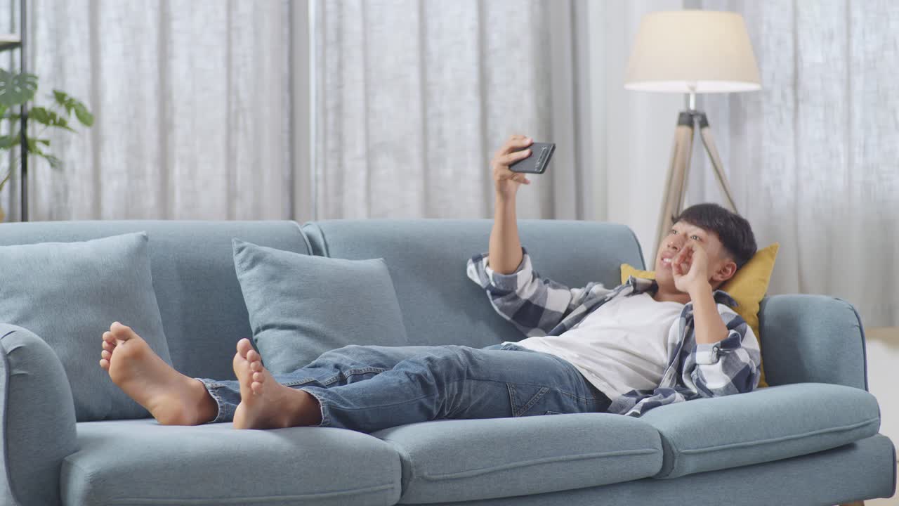 아시안 십대 소년의 몸은 집에서 거실에서 소파에 누워서 셀피를 찍는 스마트폰을 사용합니다. 미소 짓고, 평화를 보여주고, 엄지손가락을 올리고, 심장 몸짓을 합니다.