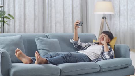 cuerpo lleno de niño adolescente asiático usando teléfono inteligente tomando selfie mientras está tendido en el sofá en la sala de estar en casa. sonriendo, mostrando paz, pulgares hacia arriba, y gesto del corazón