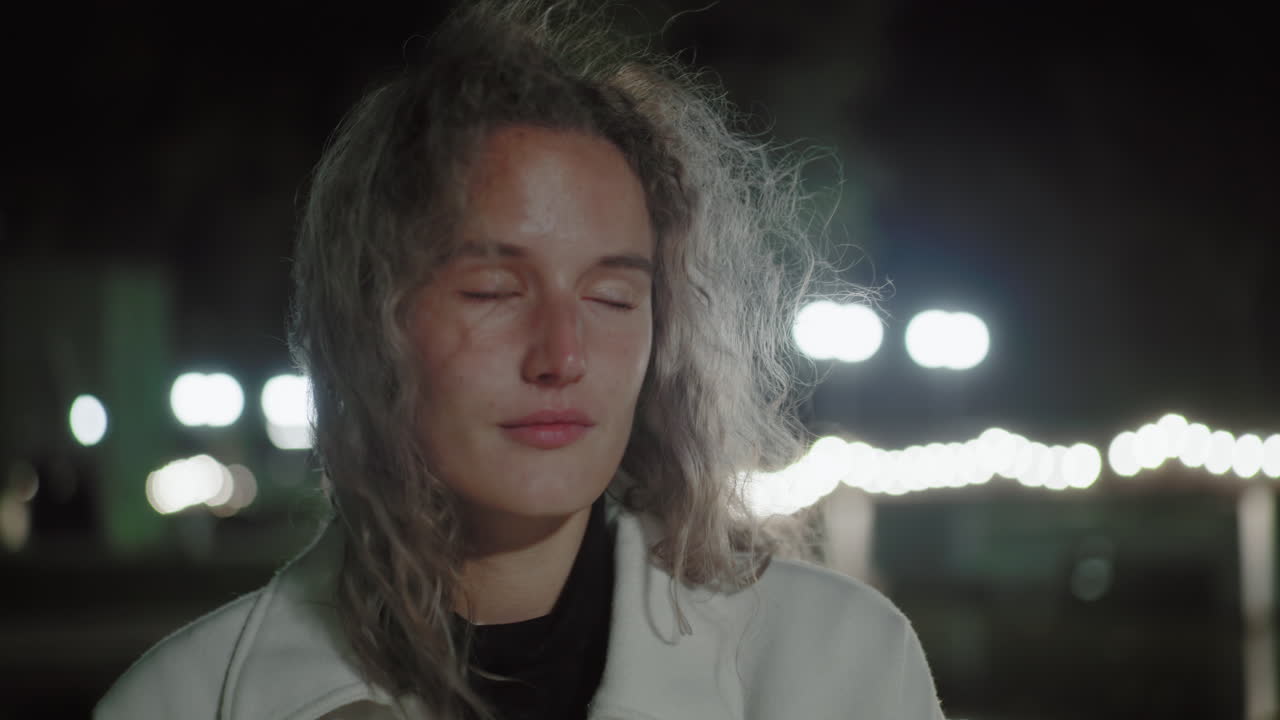 Joyful lady smiles gently alone at night with wind fluttering through her hair, blinking softly as bright streetlights blur in glowing background near calm reflective water