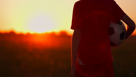 Ein-Kleiner-Fußballspieler-In-Einem-Roten-T-Shirt-Steht-Mit-Einem-Fußball-Bei-Sonnenuntergang-Auf-Dem-Feld