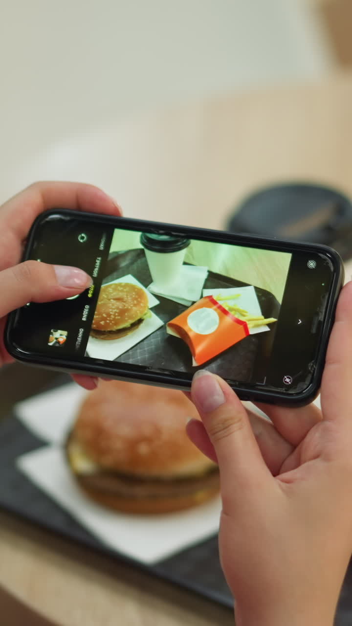 dama vista desde atrás usando su teléfono inteligente para tomar una foto de su comida, que consiste en papas fritas, una hamburguesa y una taza de café, con los alimentos dispuestos cuidadosamente en una bandeja negra en una madera