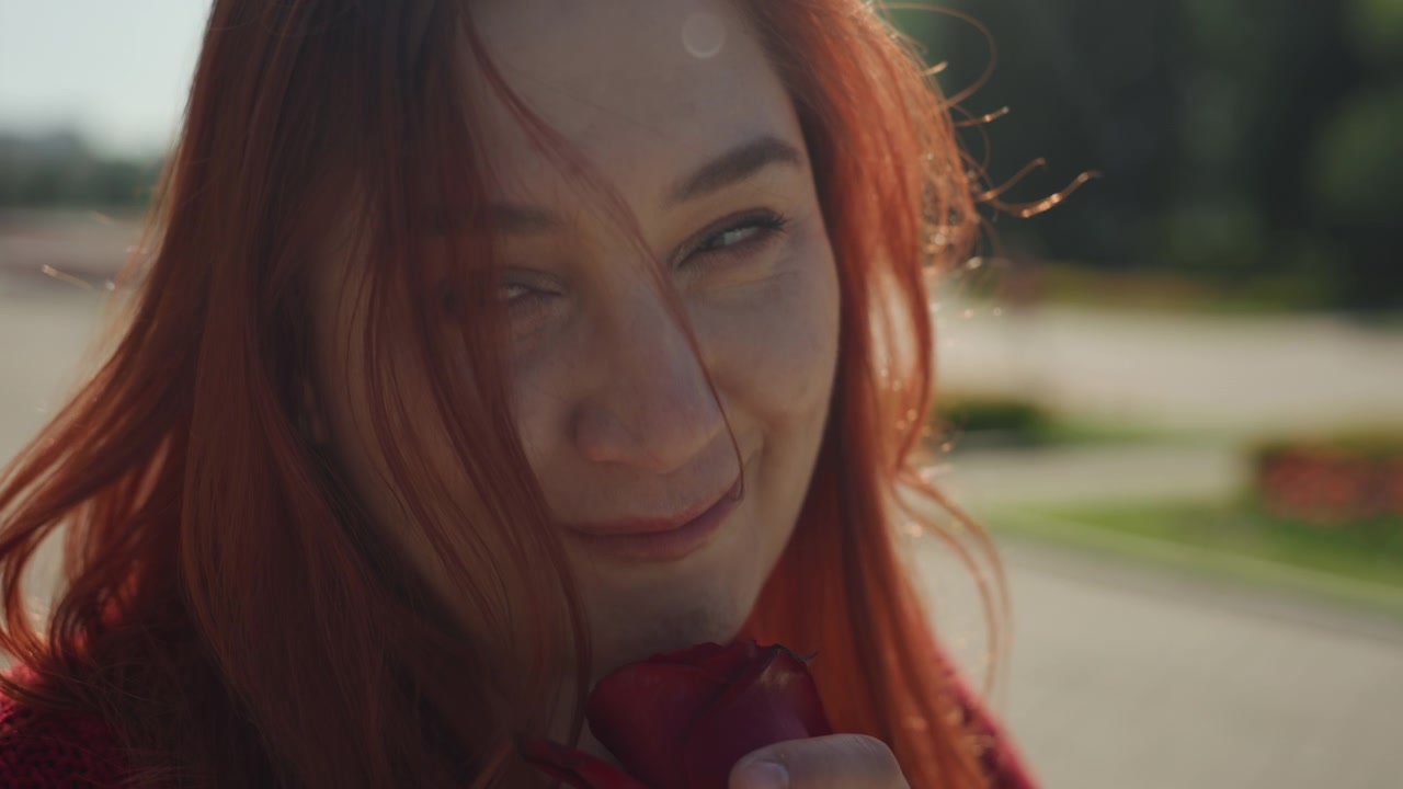 Close up of young woman smelling and holding red rose outdoors in soft natural light. Her long reddish hair flows gently as woman interacts with flower wearing textured knit sweater