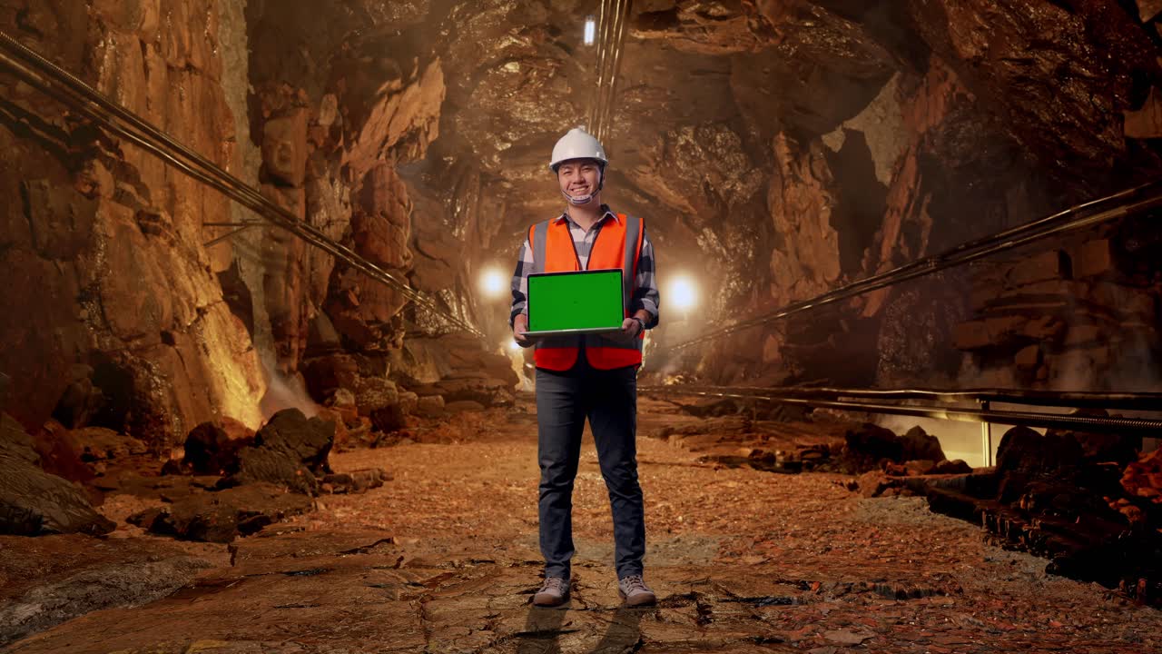 Full Body Of Asian Male Engineer With Safety Helmet Smiling And Showing Green Screen Laptop To The Camera While Standing In Underground Mine Tunnel