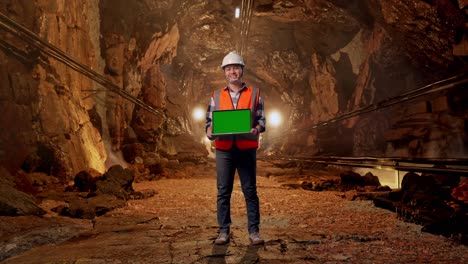 full body of asian male engineer with safety helmet smiling and showing green screen laptop to the camera while standing in underground mine tunnel