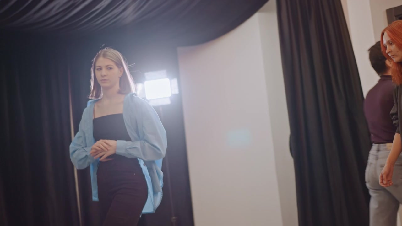 Estudiante caucásica camina por el pasillo de audiciones hacia las luces del estudio, se une a la fila con el entrenador pelirrojo y sus compañeros, con varias capas de vestuario, mostrando nerviosa anticipación, en un ensayo de casting observado por el director.