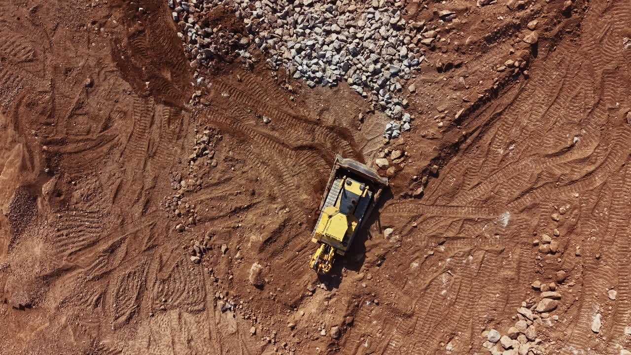 Construction equipment works on a dirt site moving rocks and soil during daylight hours near a construction area