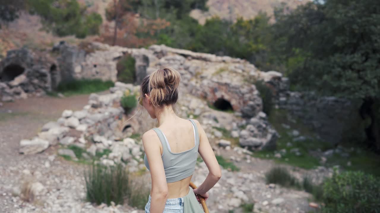 Rear view woman with wooden stick walks by rocky hills or ruins