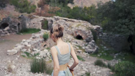 Rear-view-woman-with-wooden-stick-walks-by-rocky-hills-or-ruins