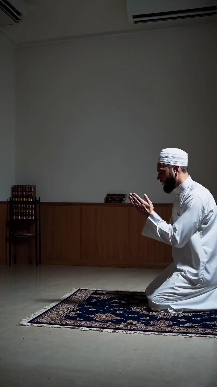 A man in traditional attire kneels in prayer on a rug, captured from a side angle
