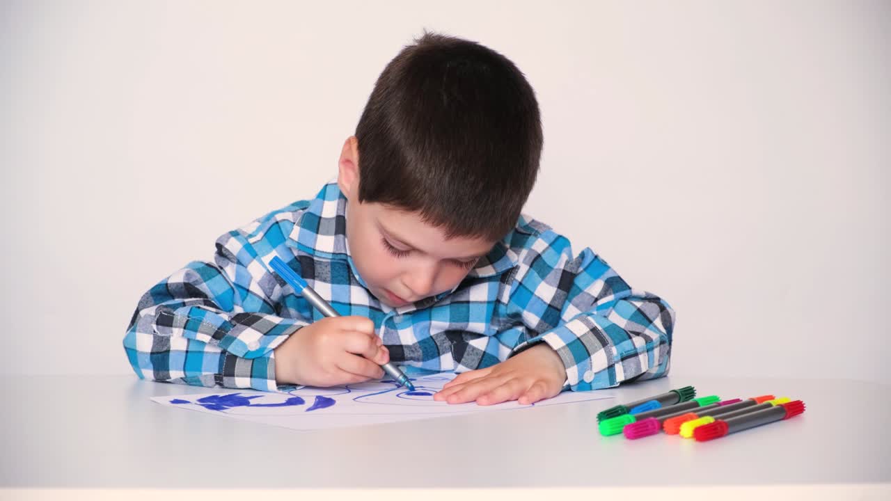 플레이드 셔츠를 입은 유치원 아이가 그림을 그리고, 파란 마커로 그림을 그립니다.