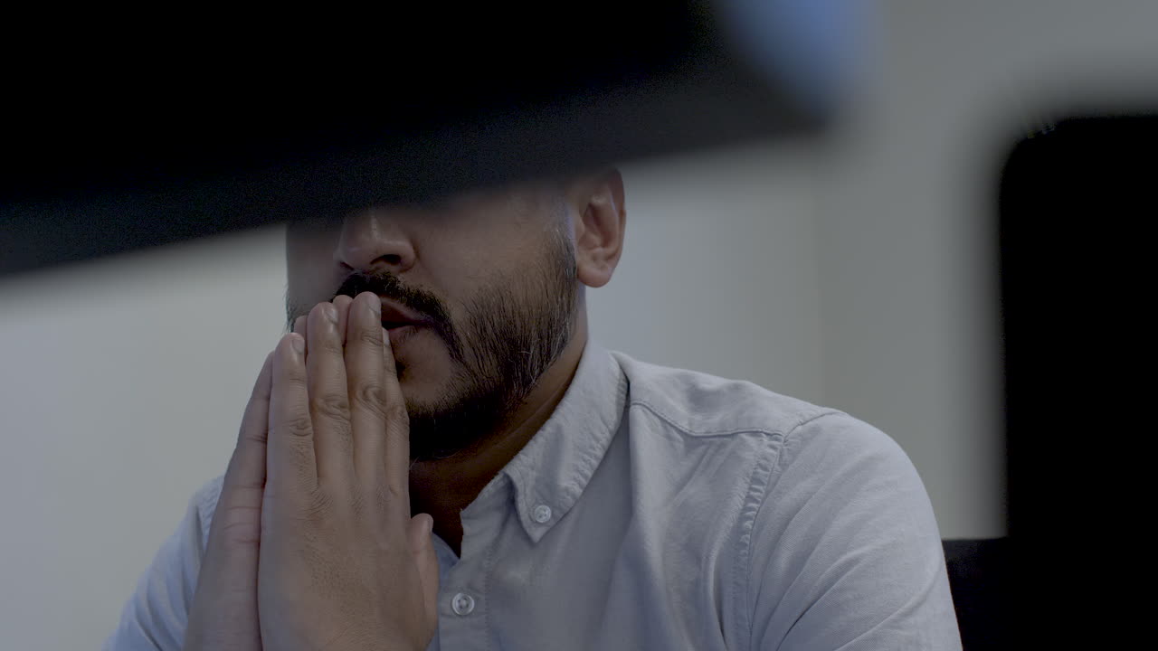 Ethnic Minority Male Office Professional Clasped Hands In Prayer, Viewed behind two computer monitors. Low Angle, Static, Shot