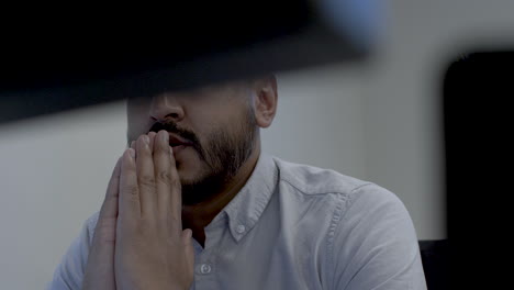 ethnic minority male office professional clasped hands in prayer, viewed behind two computer monitors. low angle, static, shot