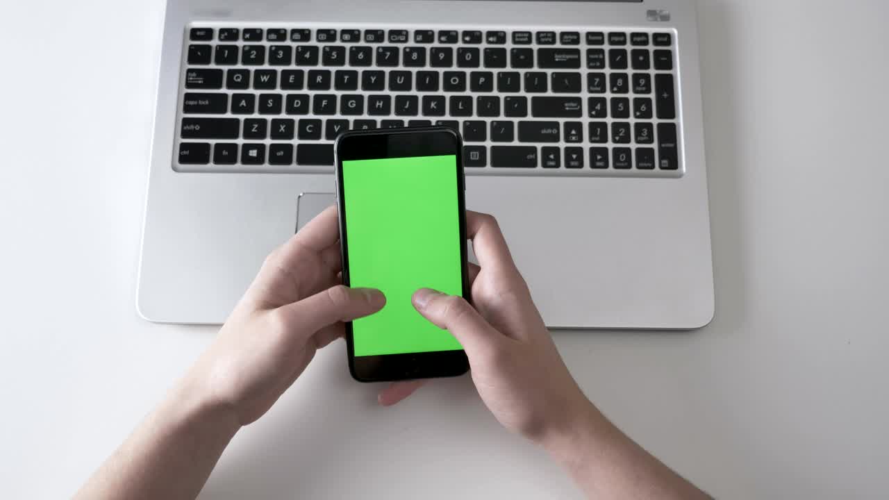Male hands typing on a larger black smartphone, laptpo in the background. Green screen, chromakey concept, laptop in the background. Top shot 60 fps