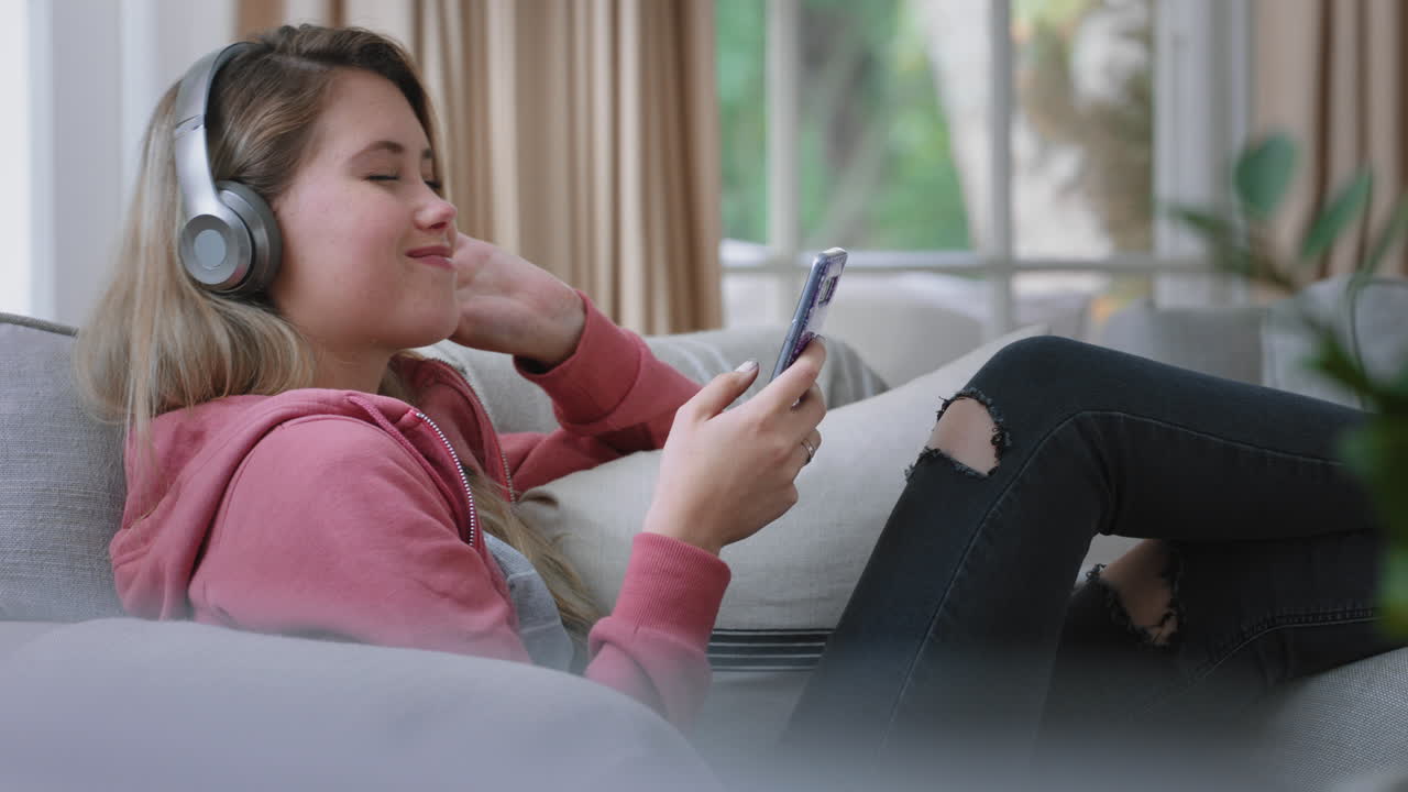 행복한 십대 소녀가 음악을 듣고 헤드폰을 착용하고 집의 소파에서 휴식을 취하고 스마트폰으로 문자 메시지를 보내고 소셜 미디어에서 라이프 스타일을 공유하는 것을 즐기고 있습니다.