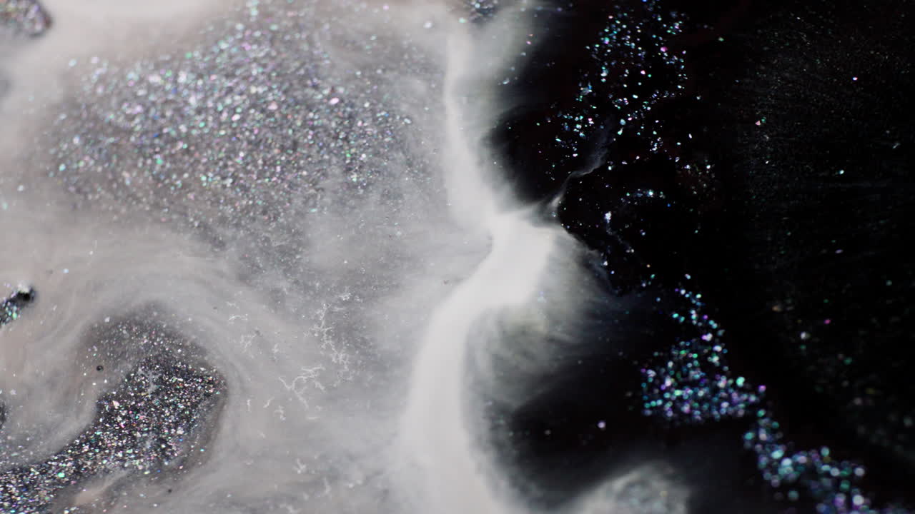 Macro view of black glitter ink dropped into a white milky fluid with silver shimmer, spreading and mixing into swirling patterns