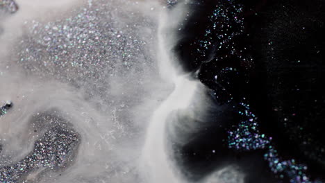 macro view of black glitter ink dropped into a white milky fluid with silver shimmer, spreading and mixing into swirling patterns