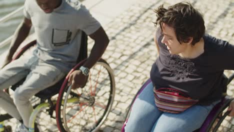 Two friends using wheelchairs wheeling at quayside and smiling