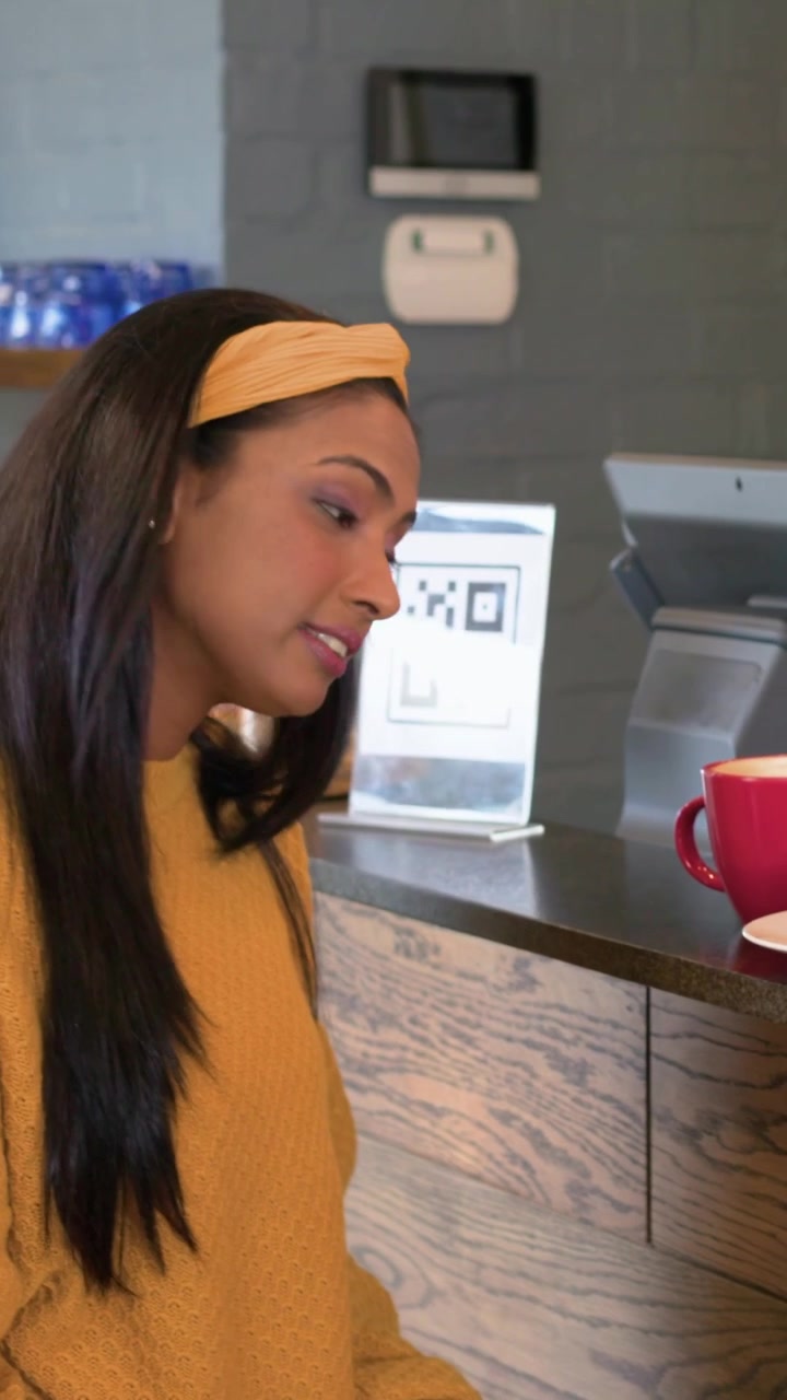 Video verticale: Una donna con un maglione color senape individua un cartello QR, appoggiata a un caffè, mentre ordina un cupcake.
