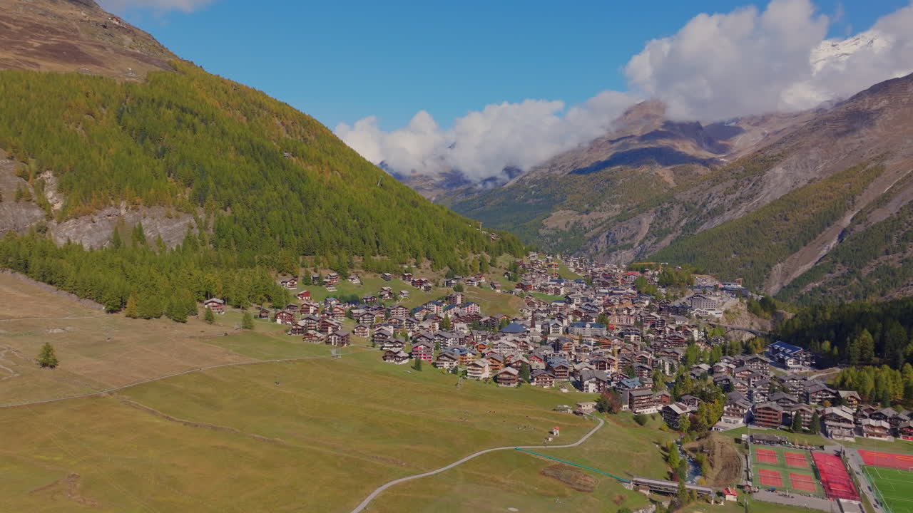 Stunning aerial footage of Saas-Fee in autumn. Golden forests surround the alpine village, nestled in the Swiss Alps. Vibrant fall colors and majestic peaks highlight the beauty of the season.
