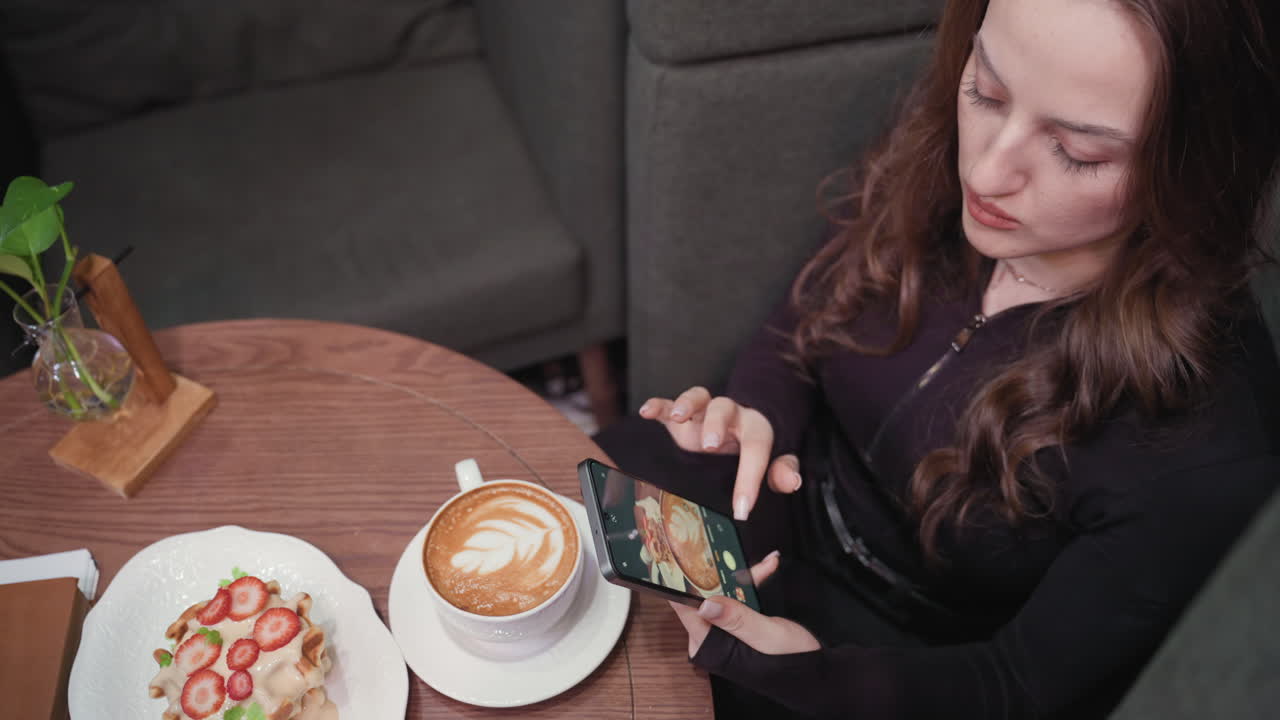 Young woman capturing overhead photo of dessert and latte using smartphone while seated in cozy green booth. Table features plant decoration, coffee art, and waffle with strawberries